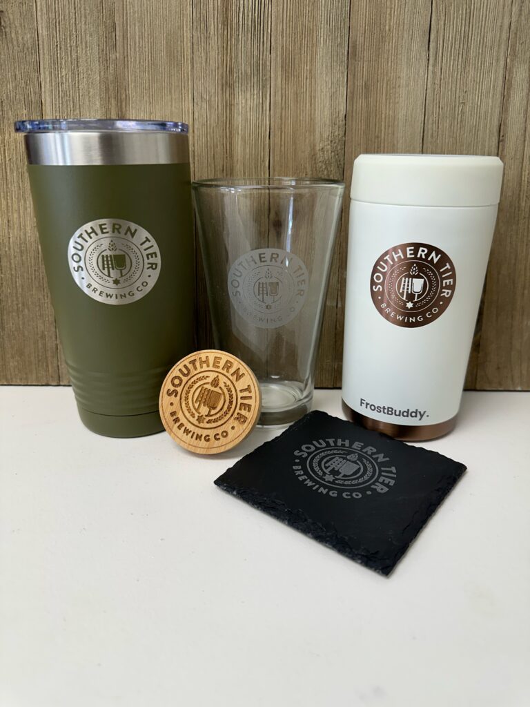 A collection of branded Southern Tier Brewing Company merchandise, including an engraved tumbler, pint glass, Frost Buddy cooler, and wooden bottle opener.