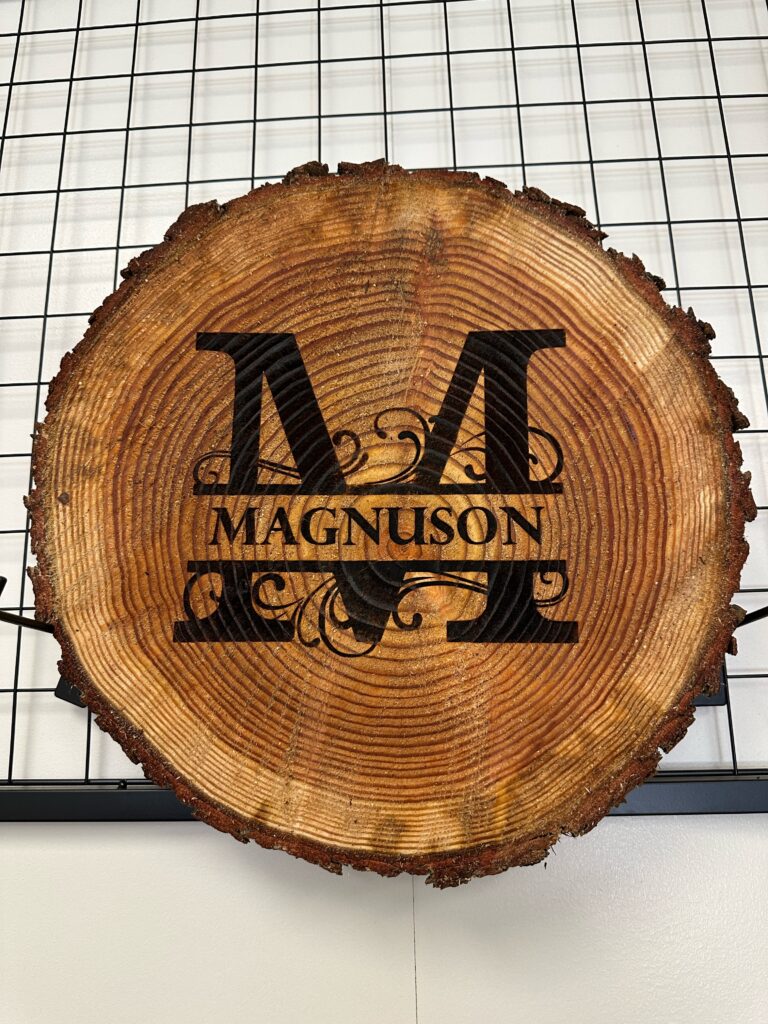 A rough-cut pine log slice with bark edge, laser-engraved with a large "M" monogram and the name "MAGNUSON"