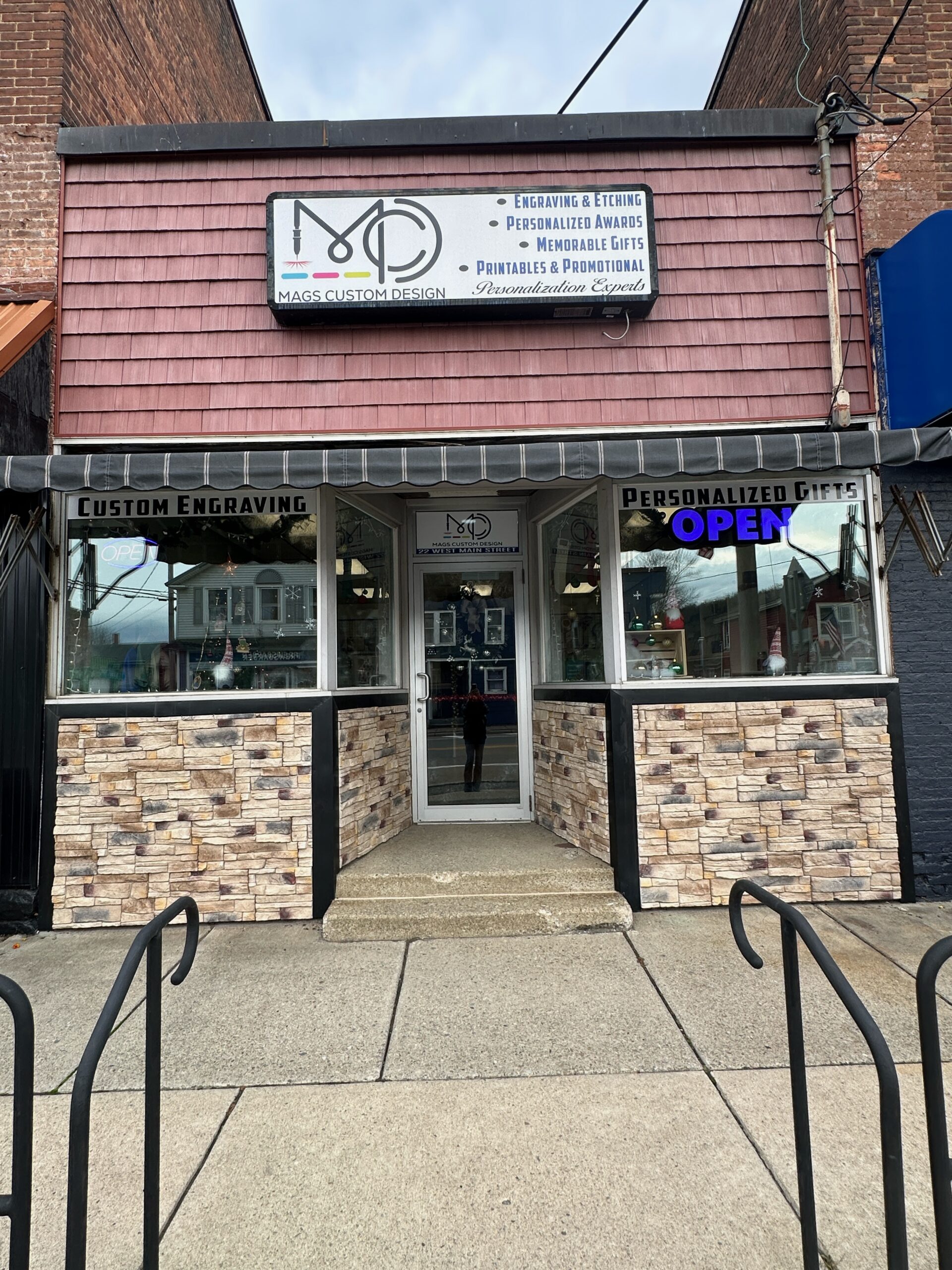 The street-level storefront of Mags Custom Design, featuring a large sign listing services like 'Engraving & Etching' and 'Personalized Gifts', with a blue 'OPEN' sign visible in the window.